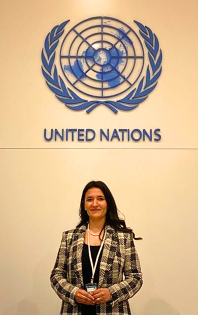 Reem El-Mograby stands in front of a wall with the United Nations logo above her head, smiling at the camera. 