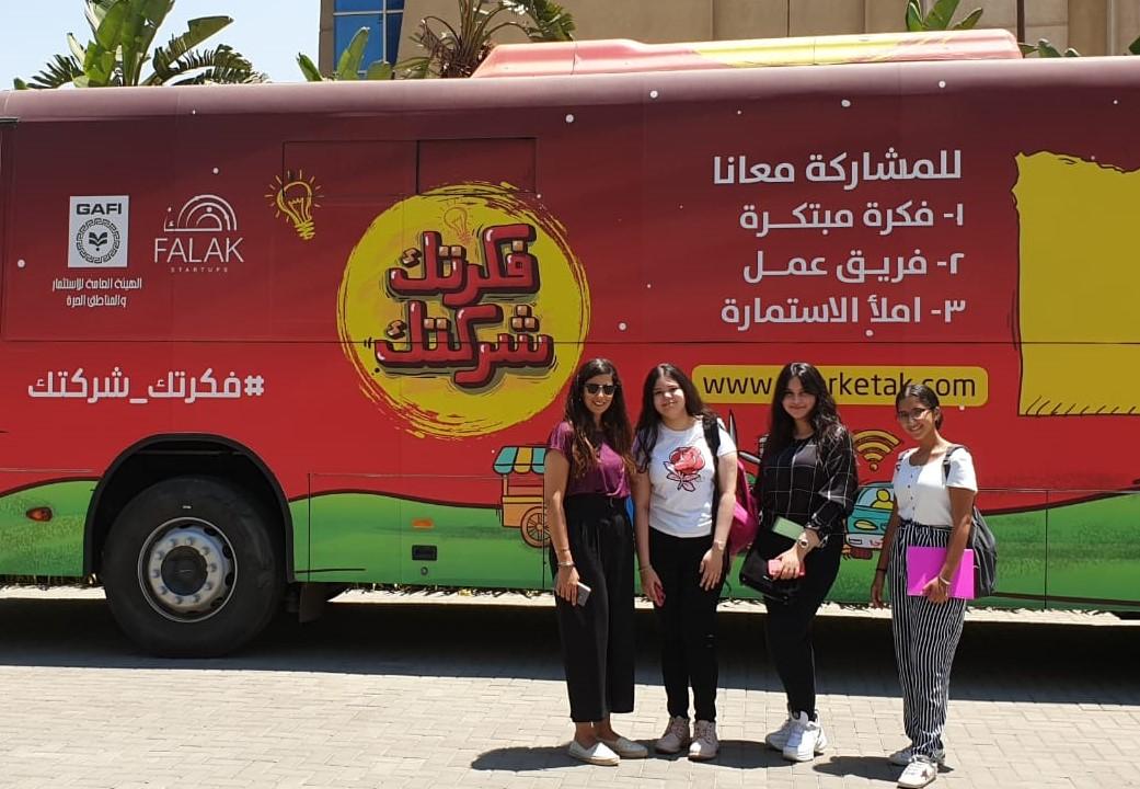 A picture of four girls in their twenties standing in front of a bus covered with texts and logos, and a main slogan that says "Your idea is Your company".
