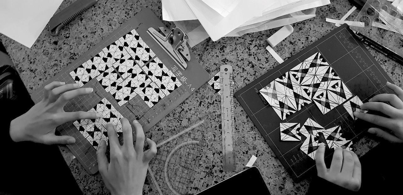 A picture of hands of two people working on intricate black and white geometric designs on clipboards, surrounded by various art supplies.