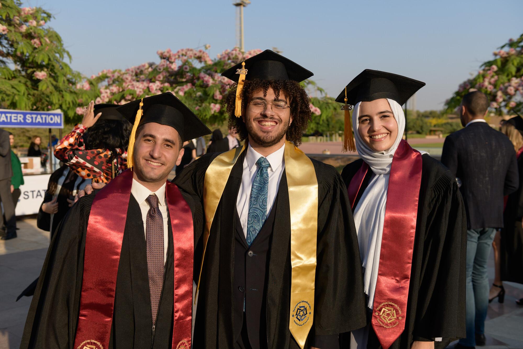 Two boys and a girl wearing a cap and gown smiling in an outdoor setting