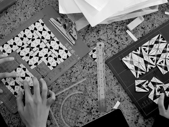 A picture of hands of two people working on intricate black and white geometric designs on clipboards, surrounded by various art supplies.