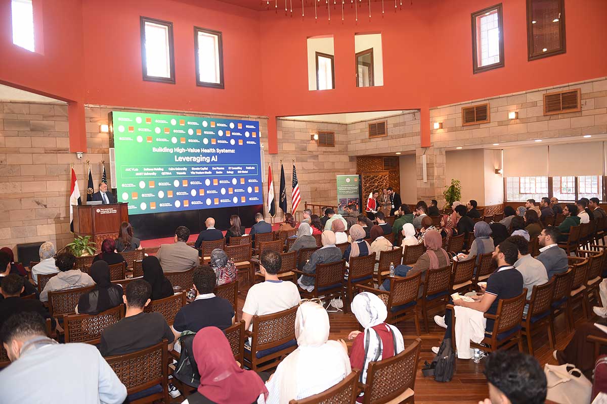 A room of seated people watch a person speak at a podium next to a large screen at the front of the room