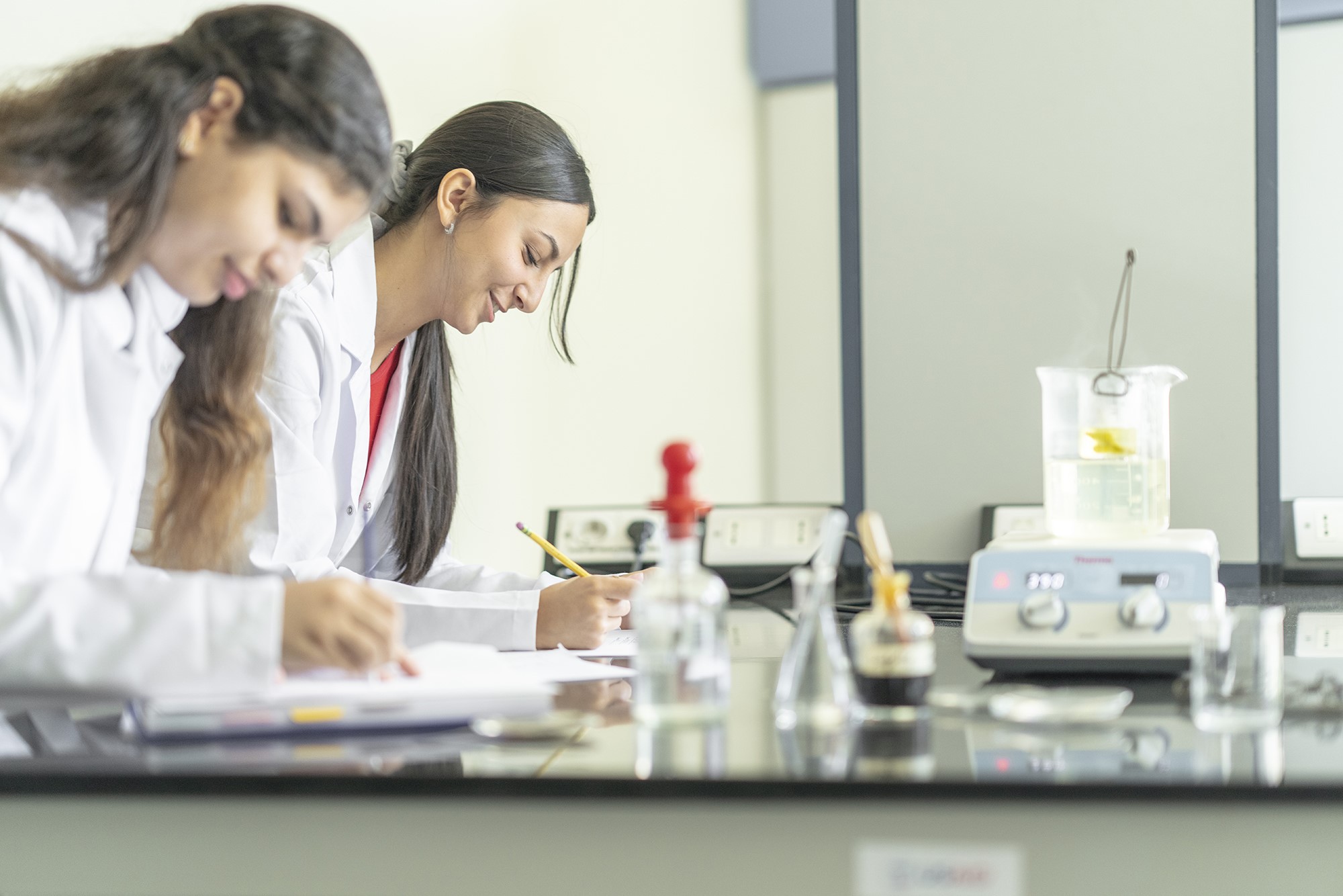 Two young women work on a counter in a science lab with glass beakers in front of them