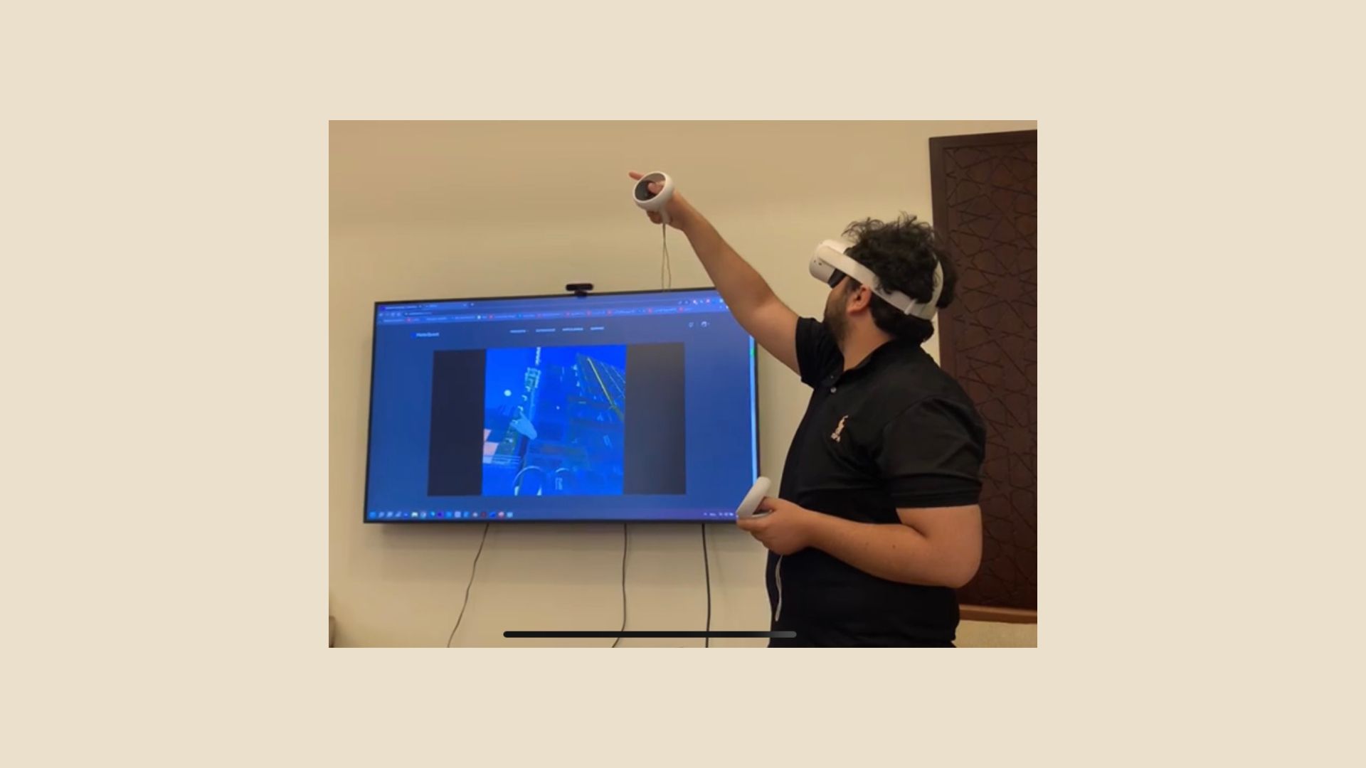 Man stands wearing a virtual reality headset and controllers while a screen behind him shows what he is seeing through the equipment