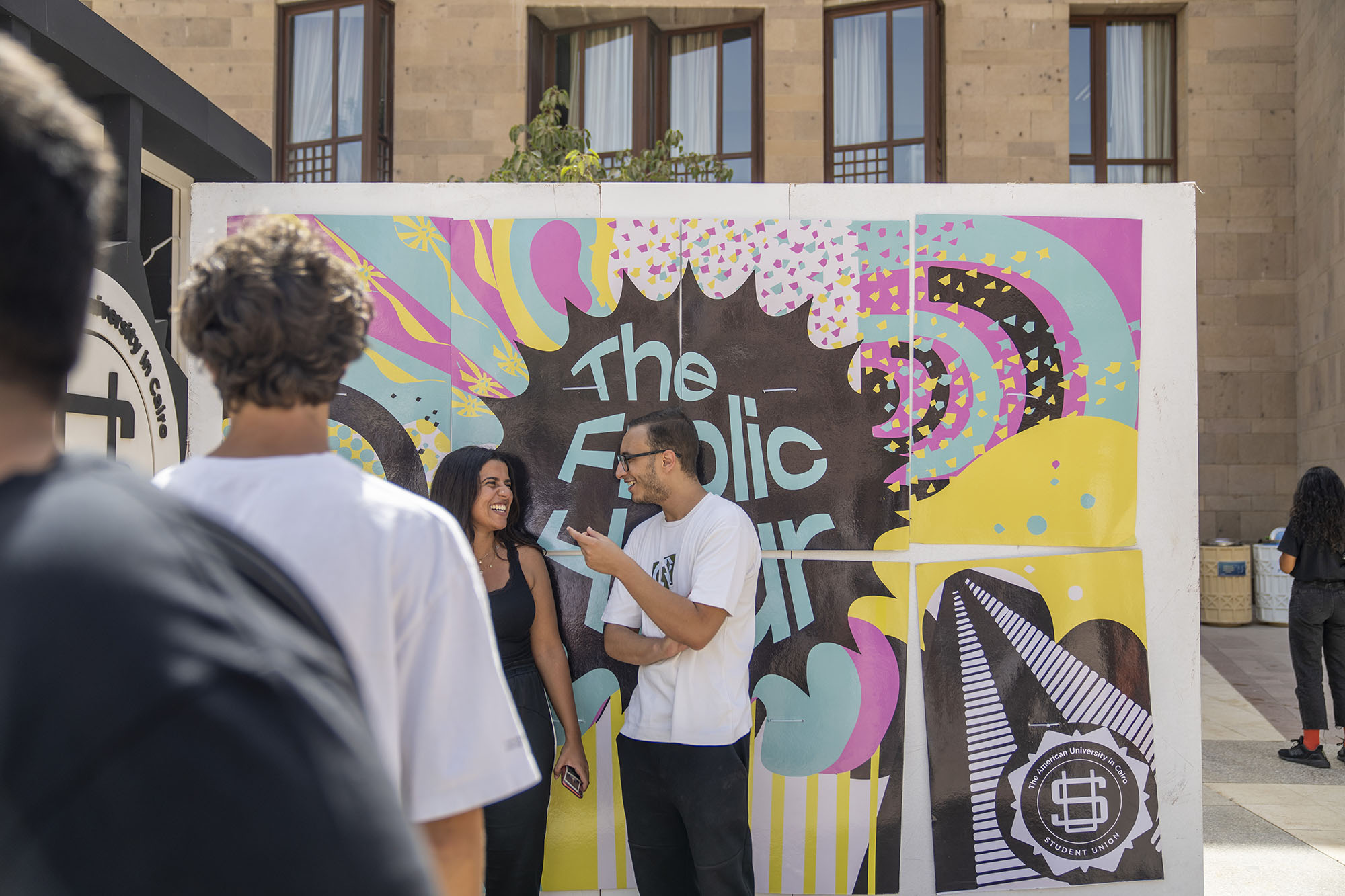 Two students standing infront of a colorful banner outdoors talking to each other