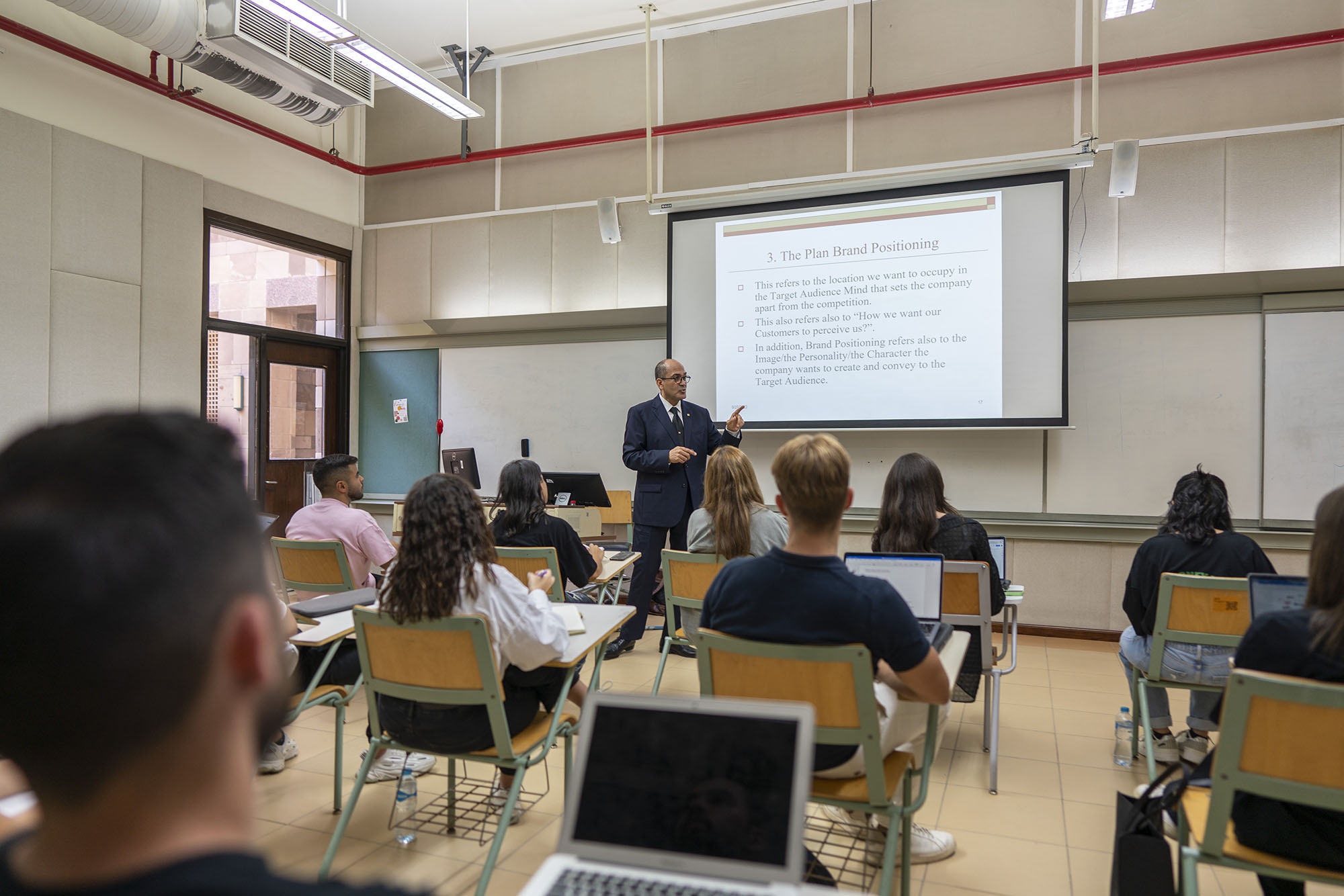 Back view of a classroom full of students with a professor giving a lecture in front of a projector