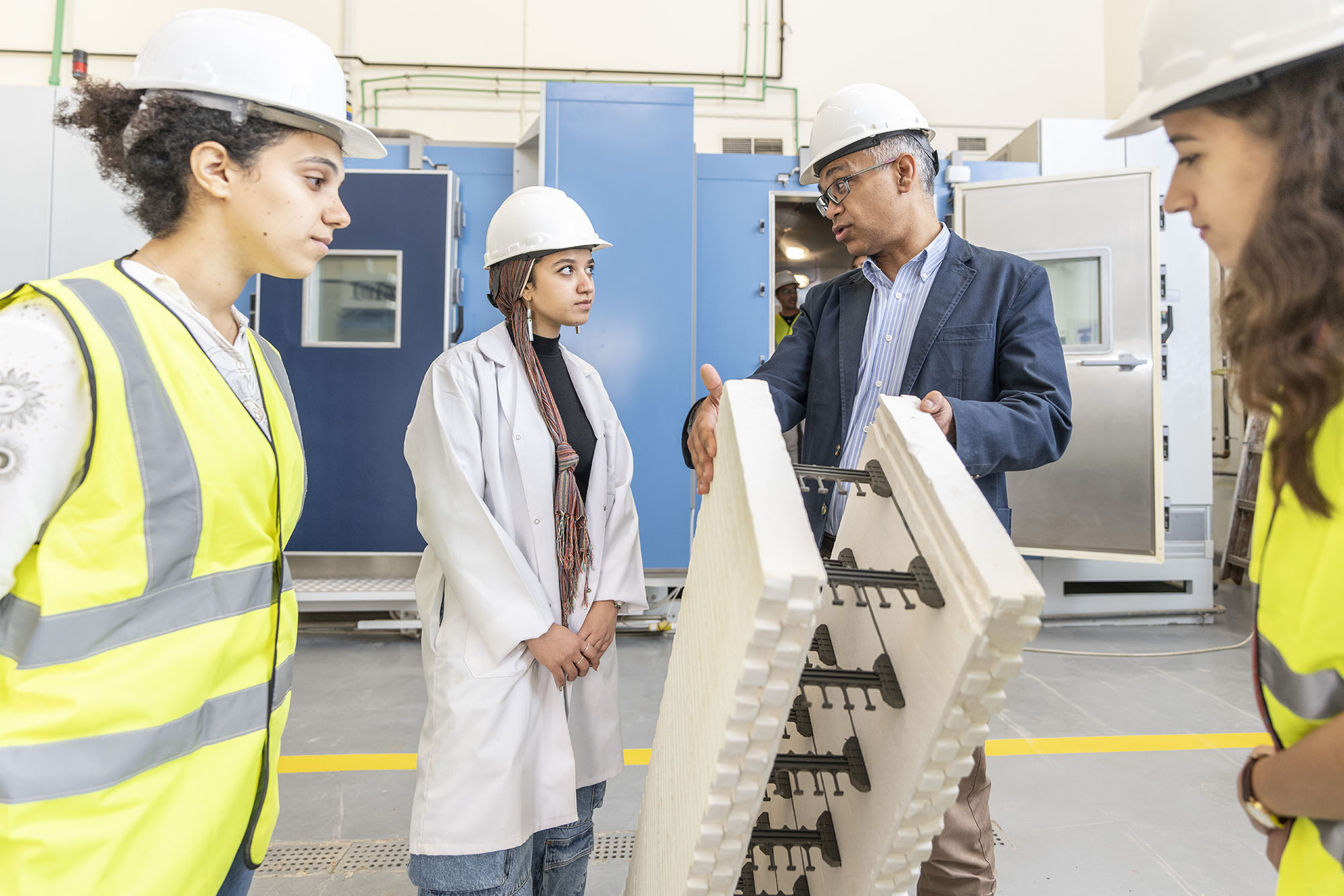 three girls wearing a yellow safety vest and helmet in a lab with a man wearing a suit explaining 