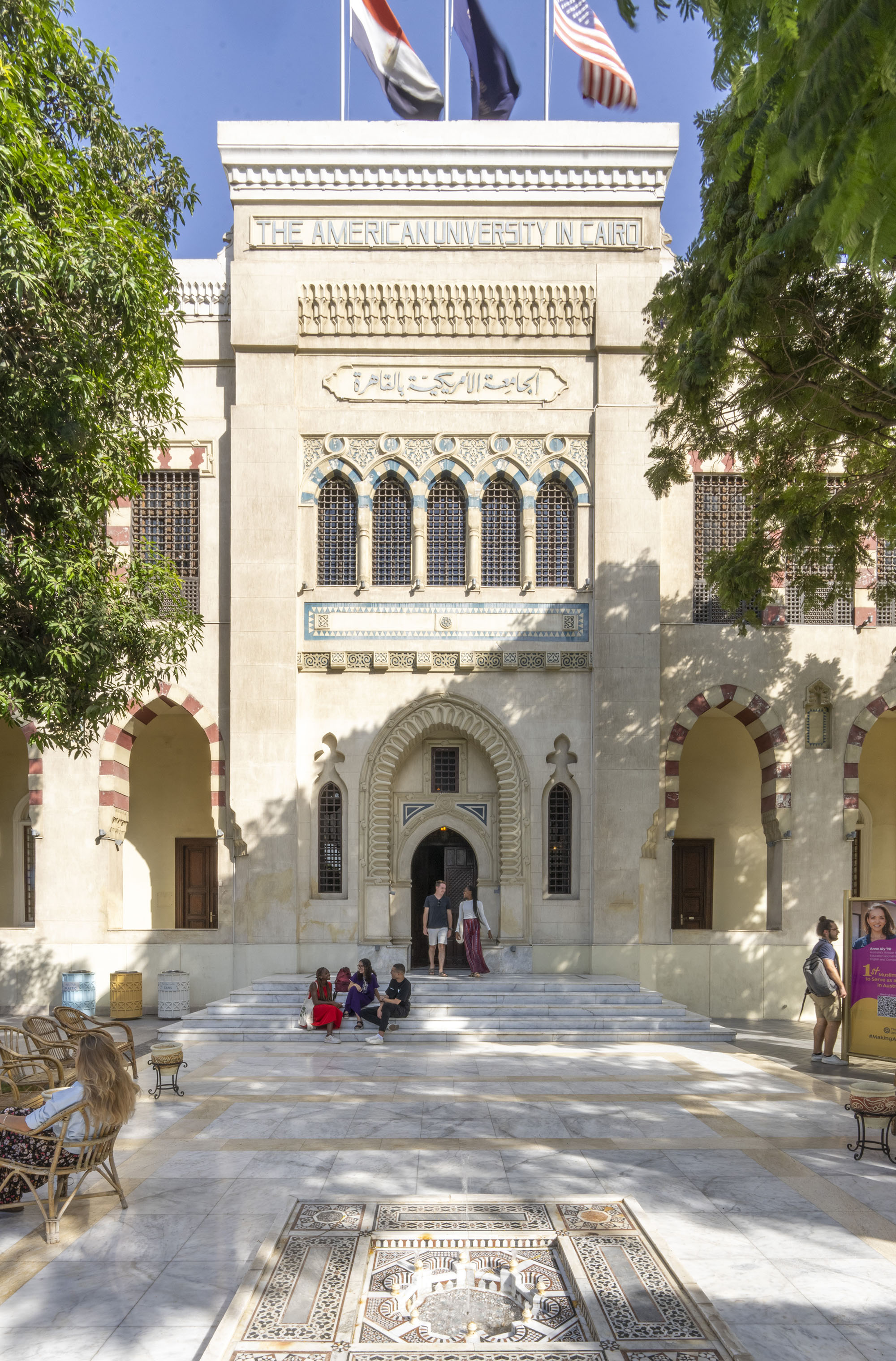 Female and male students walking on campus in front large Islamic building with three flags on the top