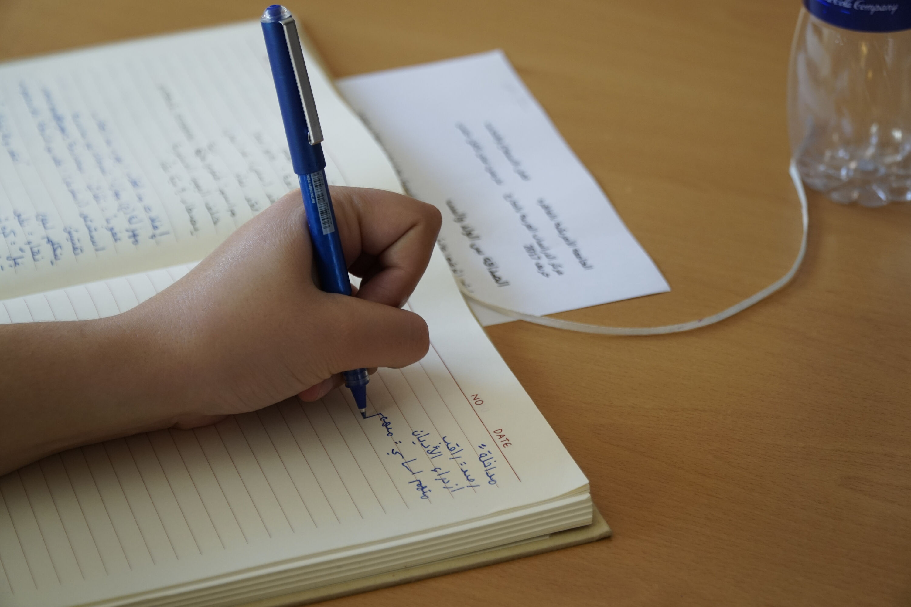 Open copybook with a hand holding a pencil on it writing in Arabic