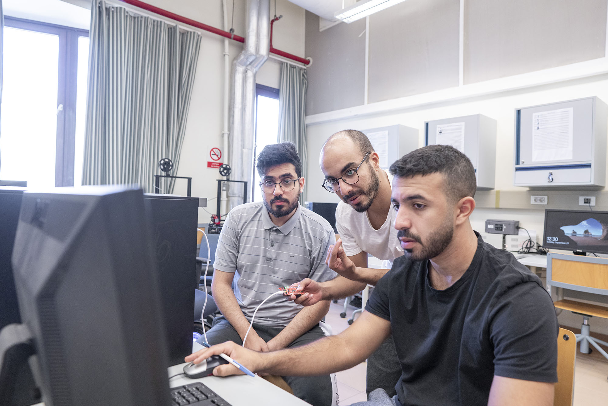 Three boys sitting on a desktop in a computer lab