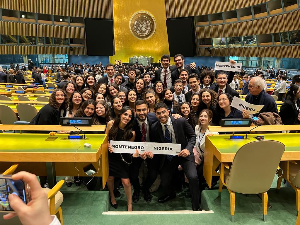 The CIMUN team stands wearing suits and professional dresses in an auditorium at the conference. Some students are holding signs saying "Montenegro" and "Nigeria"