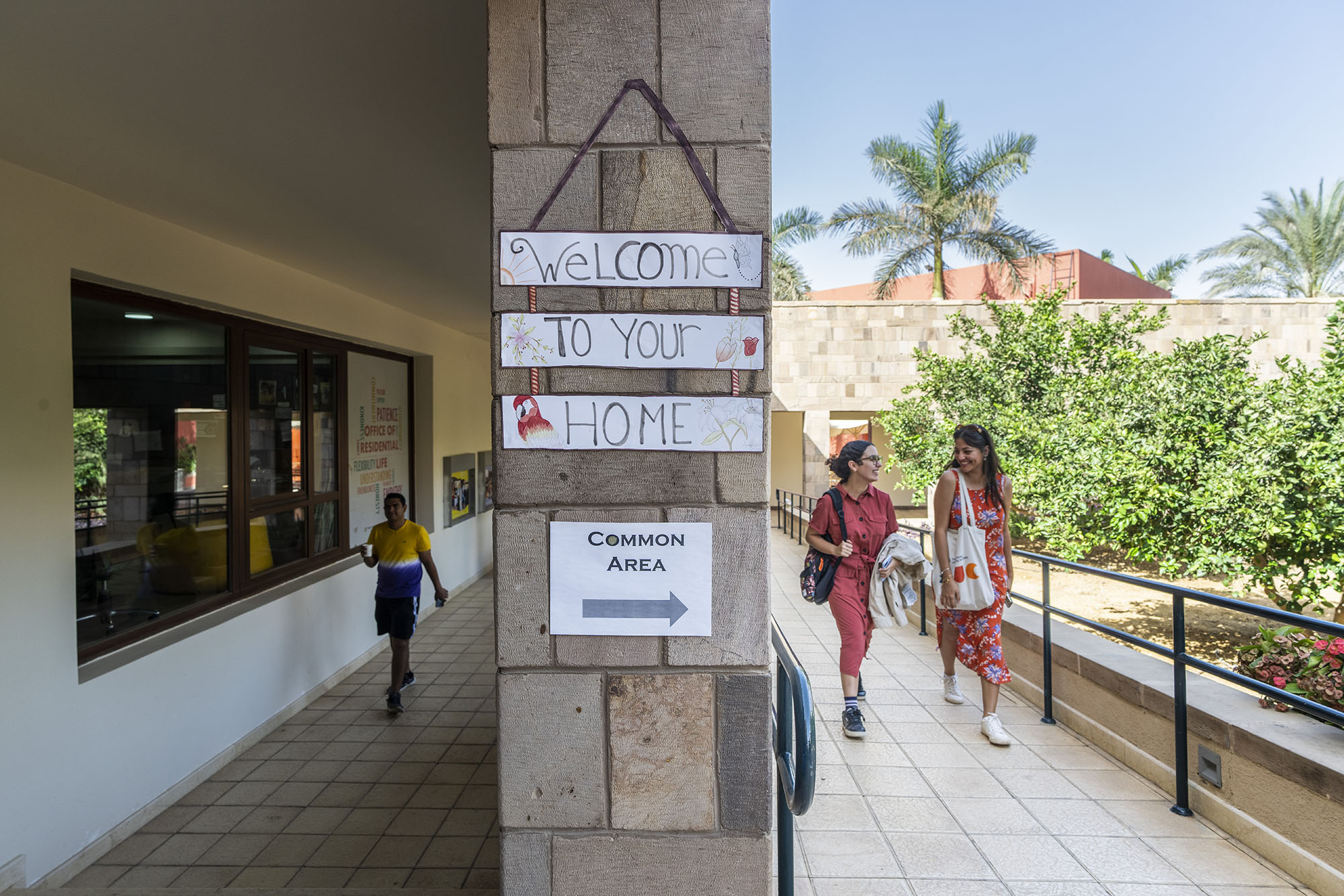 Two female students walking and talking together and a male student walking and holding a drinking cup, igns "Welcome to your home" and "Common area"