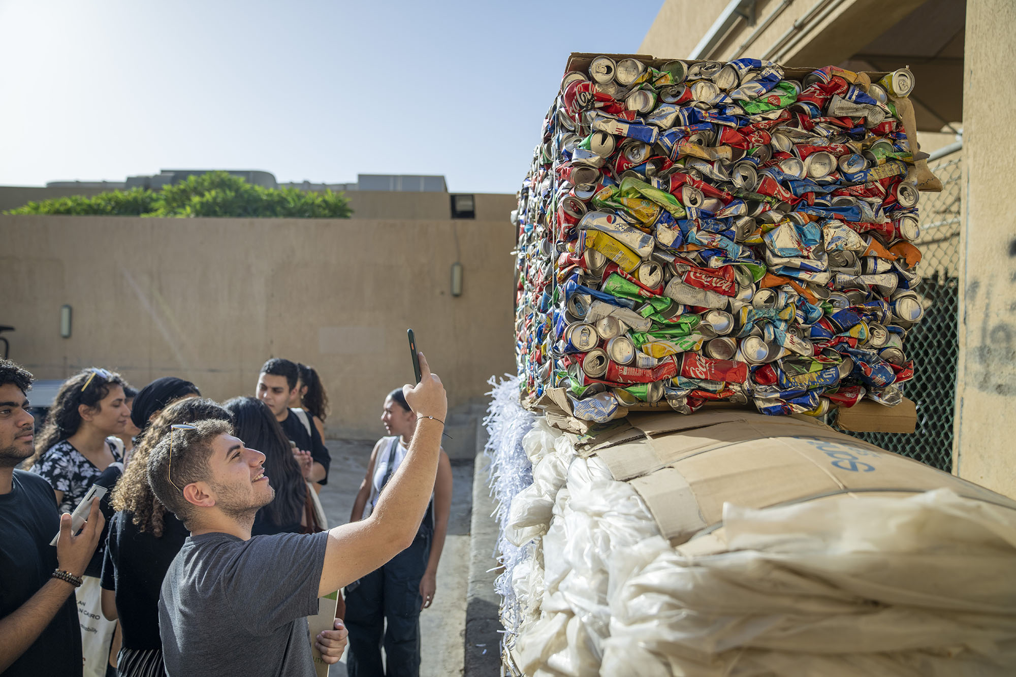 student taking a photo with his phone of the large pile of recycling packed soft drinks pile ready for recyling