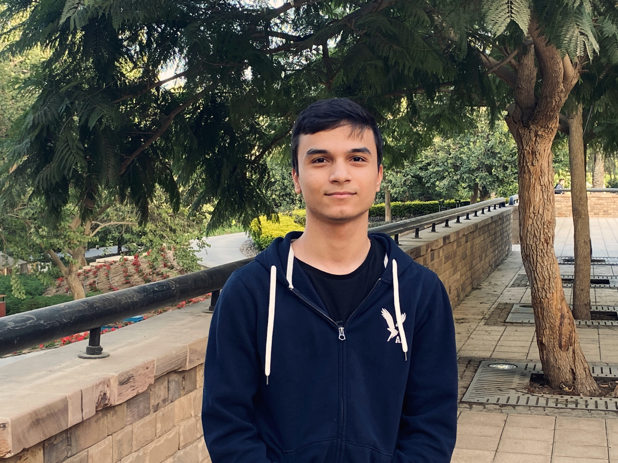 Young man with short dark hair stands in front of trees on a college campus, wearing a navy hoodie