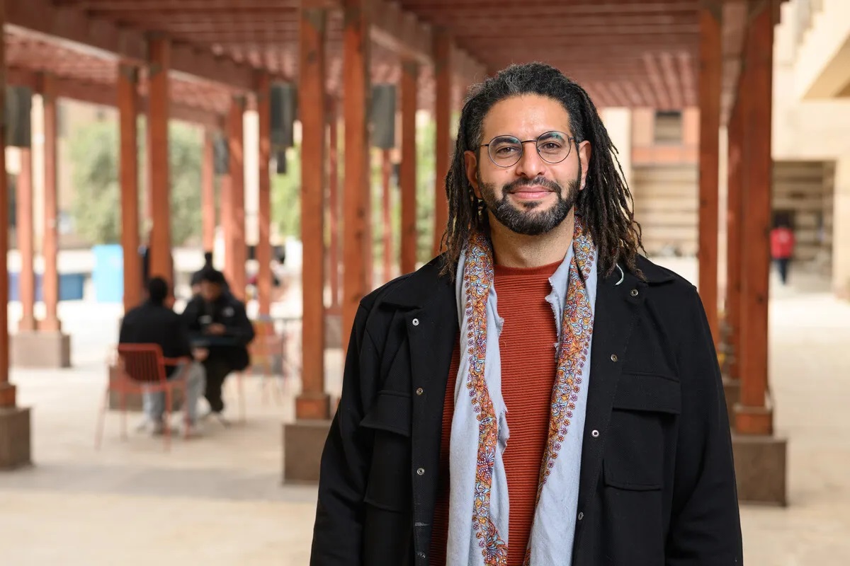 A bearded man is smiling and wearing glasses is standing outdoors
