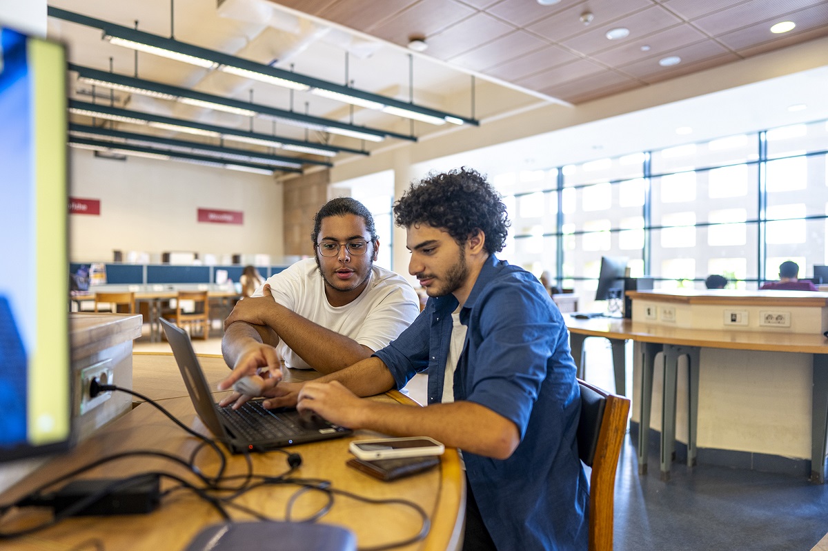 Two males are sitting at a desk in front of a laptop in a classroom