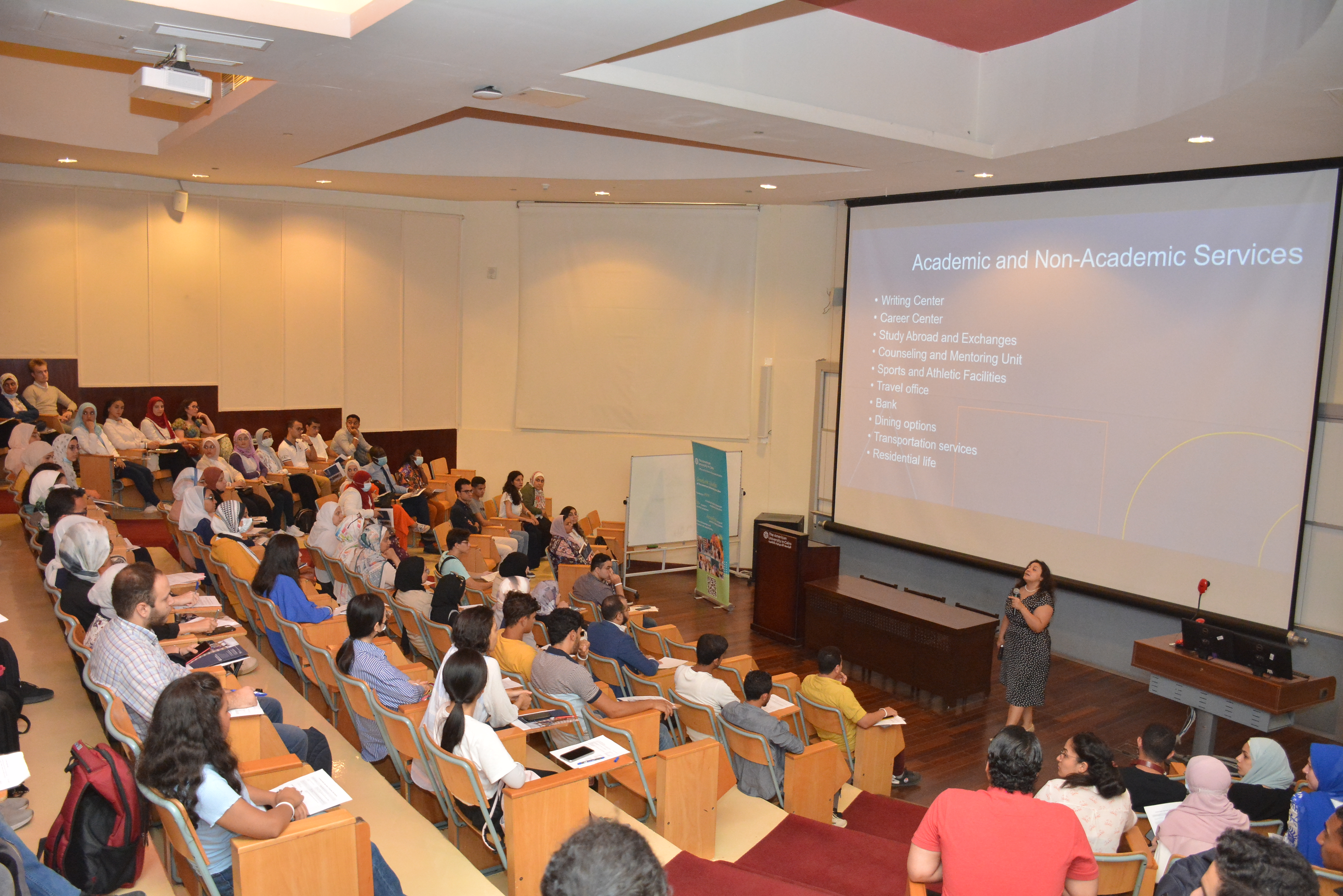 A woman is giving a presentation in a large lecture hall, where many students are seated and listening. A big screen behind her shows a list of academic and non-academic services