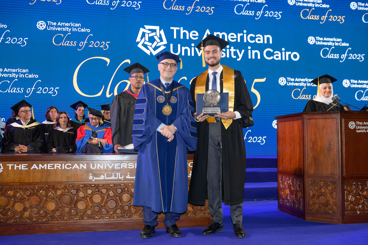 Commencement stage at The American University in Cairo, with a graduate proudly holding an award next to AUC President in blue robe, as other professors and officials look on