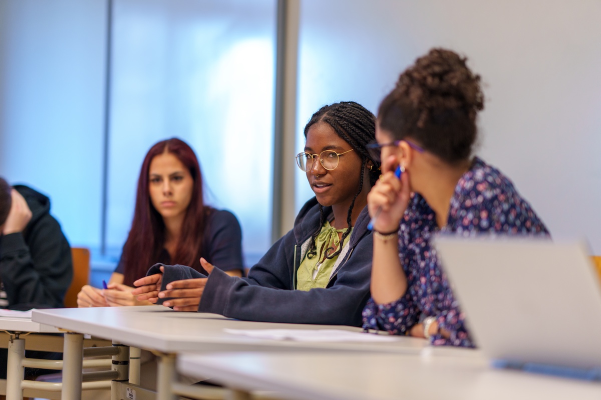 A university student with braided hair and glasses speaks during a classroom discussion while classmates listen around a table.