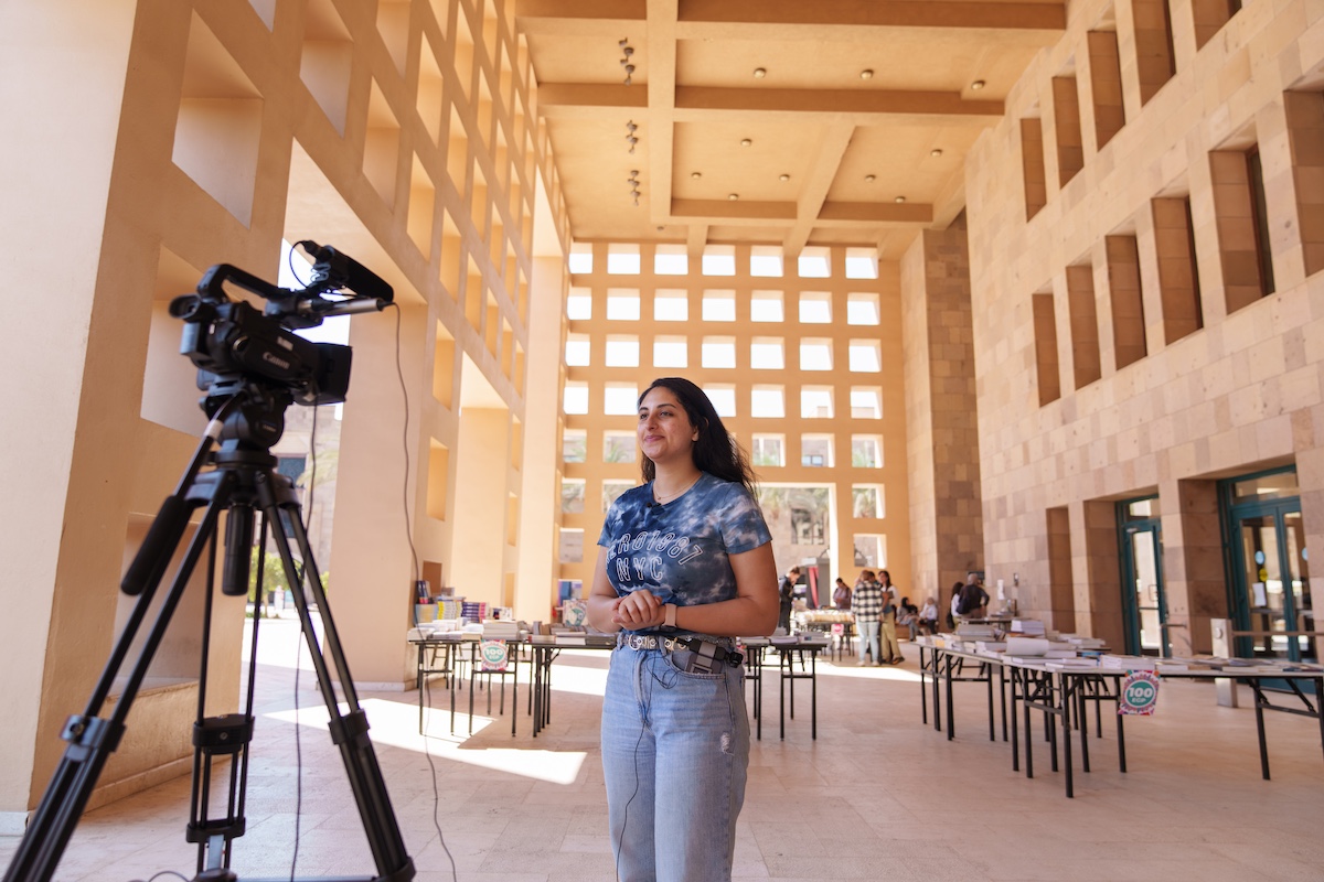 A student stands on campus speaking to a camera set on a tripod, with tables and palm trees in the background.