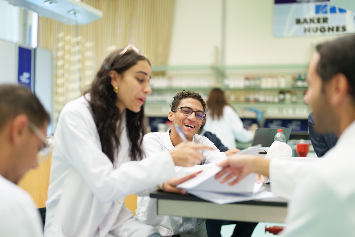 Students in white lab coats review papers together at a laboratory table, engaged in conversation.