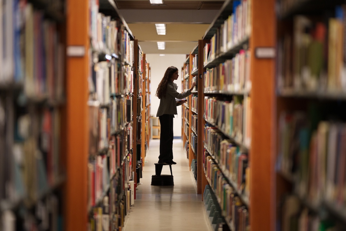A student stands on a small step stool between two library shelves while selecting a book.