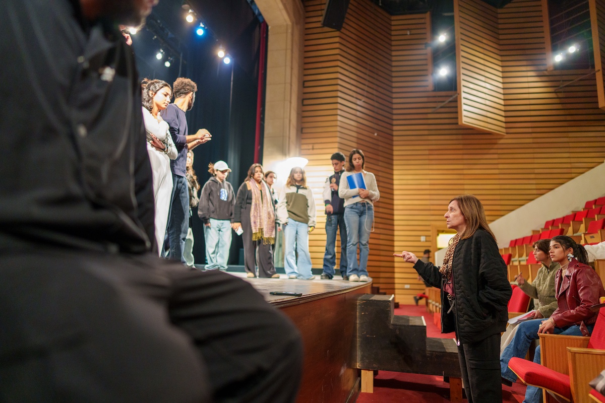 A group of students stand on a theater stage while an instructor gives feedback from the audience seating.