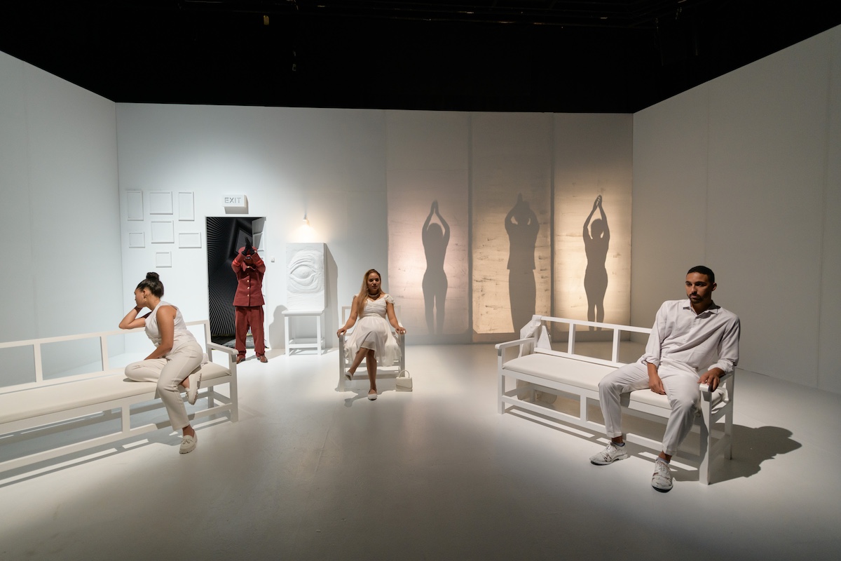 An art installation featuring people dressed in white seated on benches on a theatre with dramatic shadows cast on the walls.