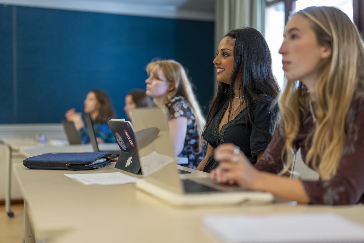 Students sit in a seminar-style classroom with laptops open.