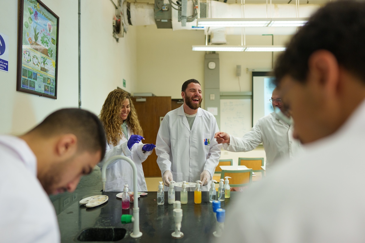 Students in lab coats laugh together around a laboratory bench with small bottles and experiment materials arranged in front of them.