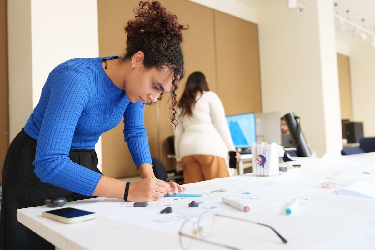 A student leans over a large table sketching and working on a design project in a studio classroom.