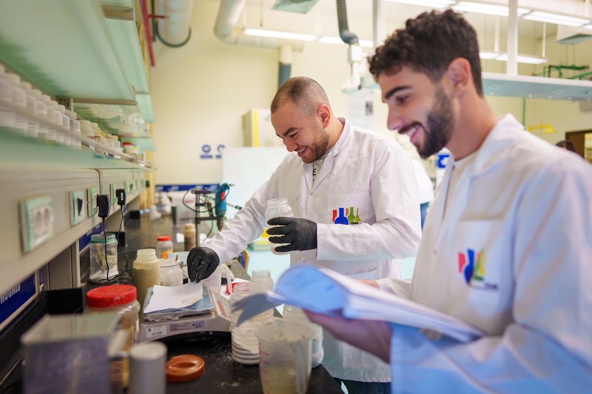 Two students wearing lab coats conduct an experiment in a laboratory while reviewing notes.