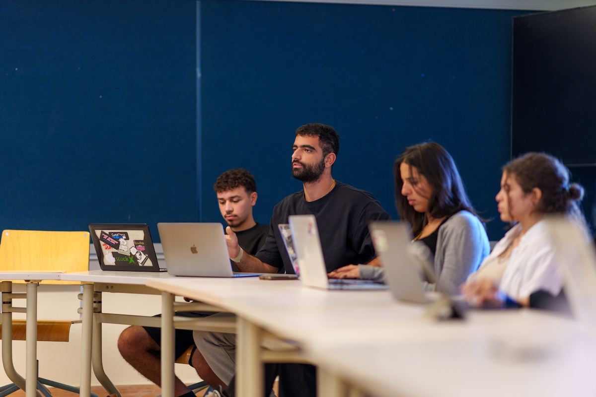 Students sit in a seminar-style classroom with laptops open.