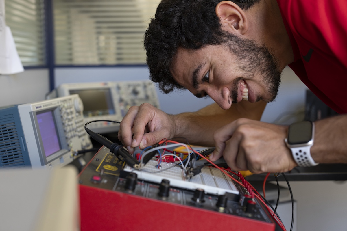A smiling student leans over an electronics workstation, adjusting wires on a circuit board connected to testing equipment.