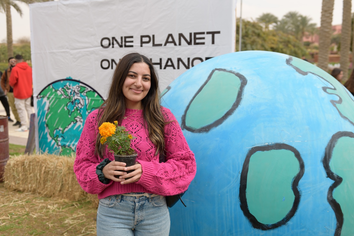 A student holds a small potted flower in front of a large Earth display and a sign reading &ldquo;One Planet, One Chance.&rdquo;