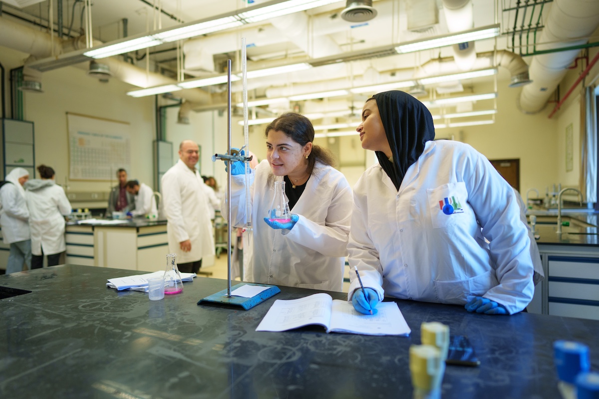 Two students in lab coats conduct a chemistry experiment using flasks and lab equipment while taking notes.