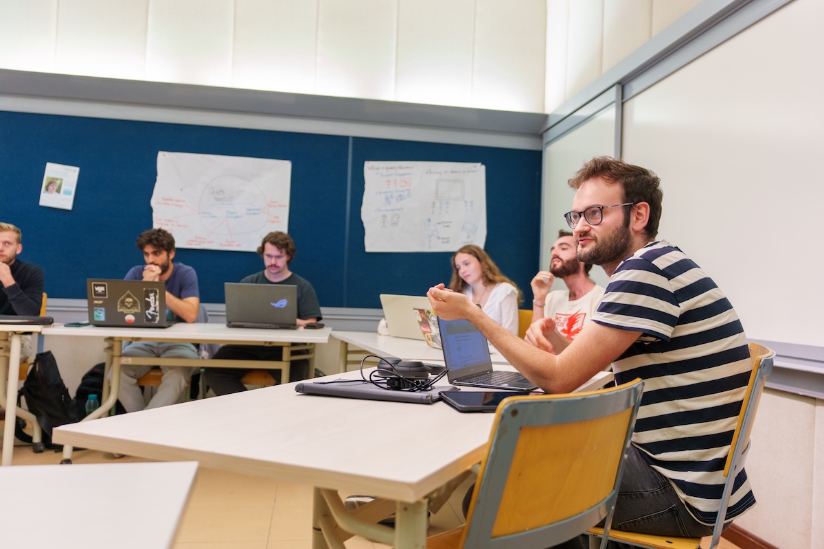 Students participate in a classroom discussion with laptops open and presentation posters displayed on the wall.
