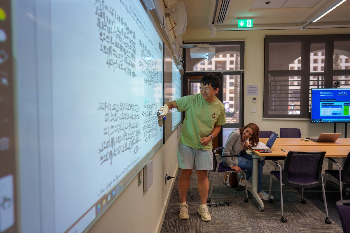 A student writes ancient Egyptian text on a digital classroom board while another student watches from a nearby table.