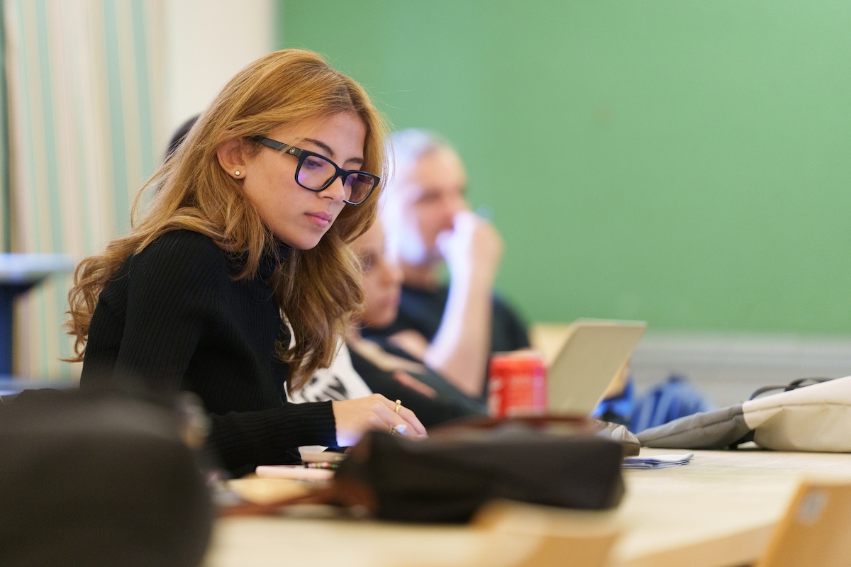 A student wearing glasses concentrates while working on a laptop in a classroom.