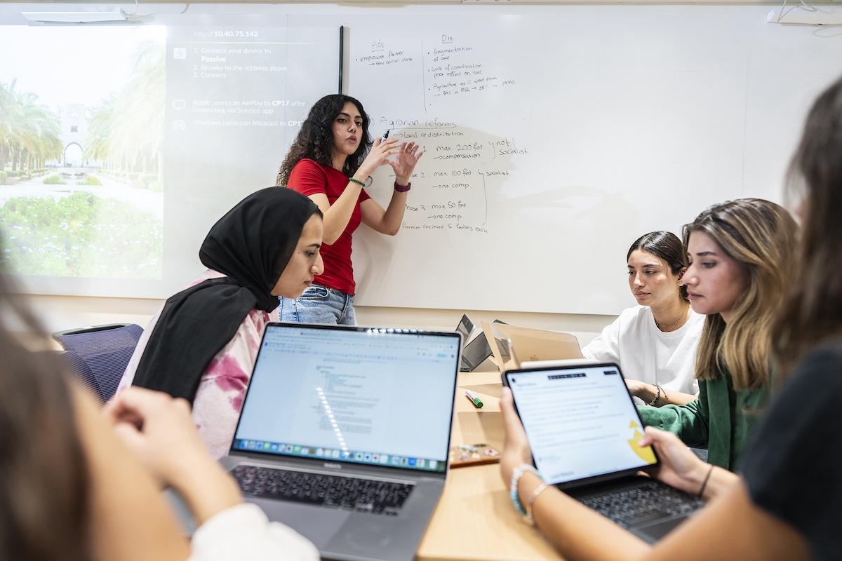 A student presents ideas at a whiteboard while classmates use laptops and tablets during a group session.
