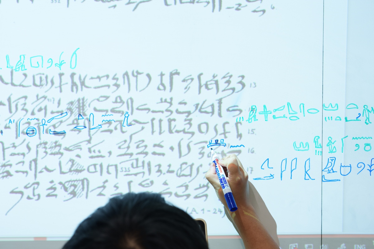 Close-up of a hand writing symbols and notes in blue marker on a whiteboard filled with layered writing and diagrams.