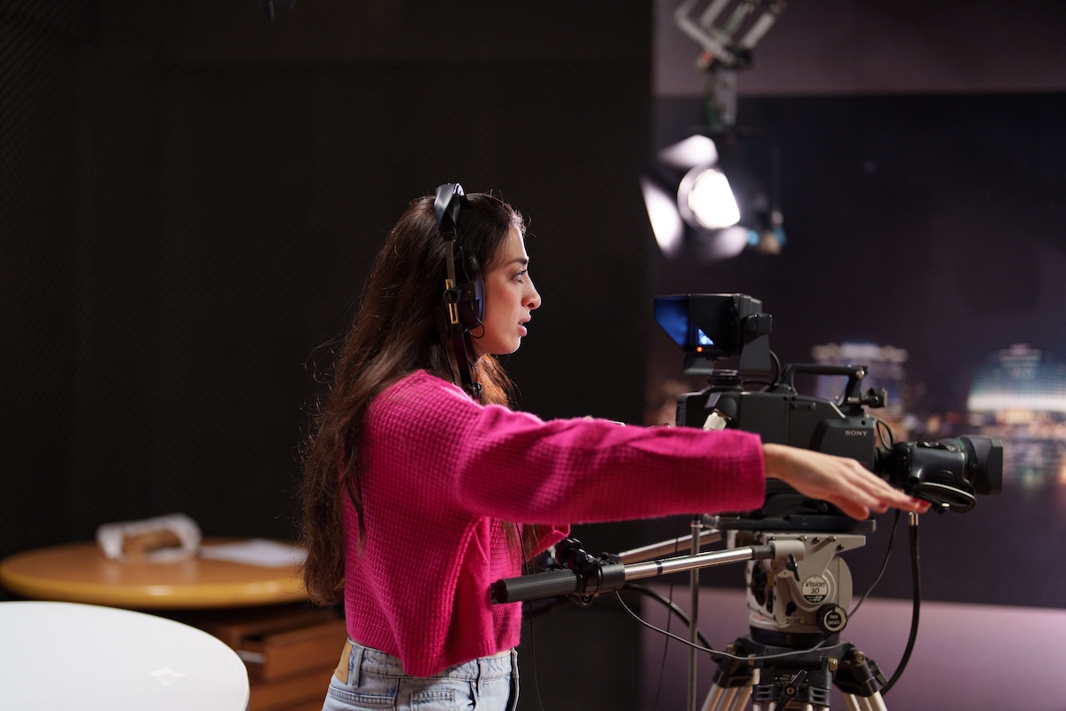 A student wearing headphones operates a professional video camera on a tripod in a studio setting.
