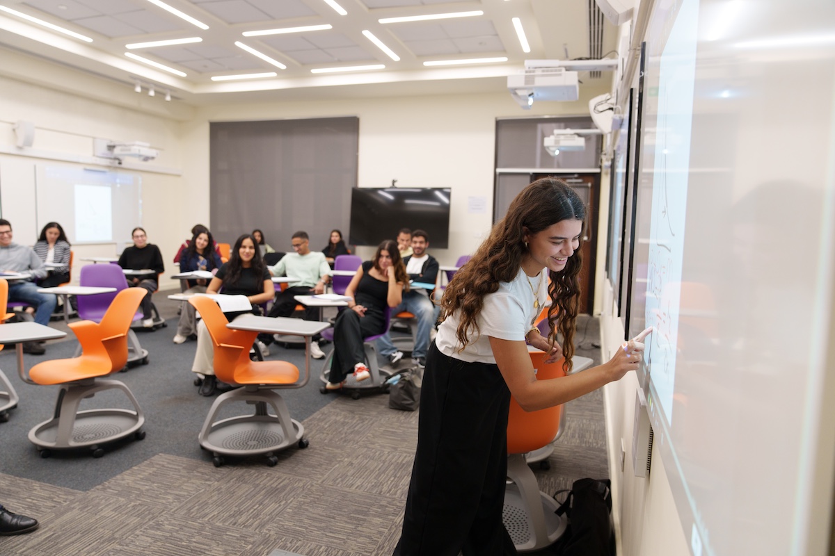 A student smiles while writing on a digital classroom board as classmates sit behind her in a interactive style classroom with colorful chairs.