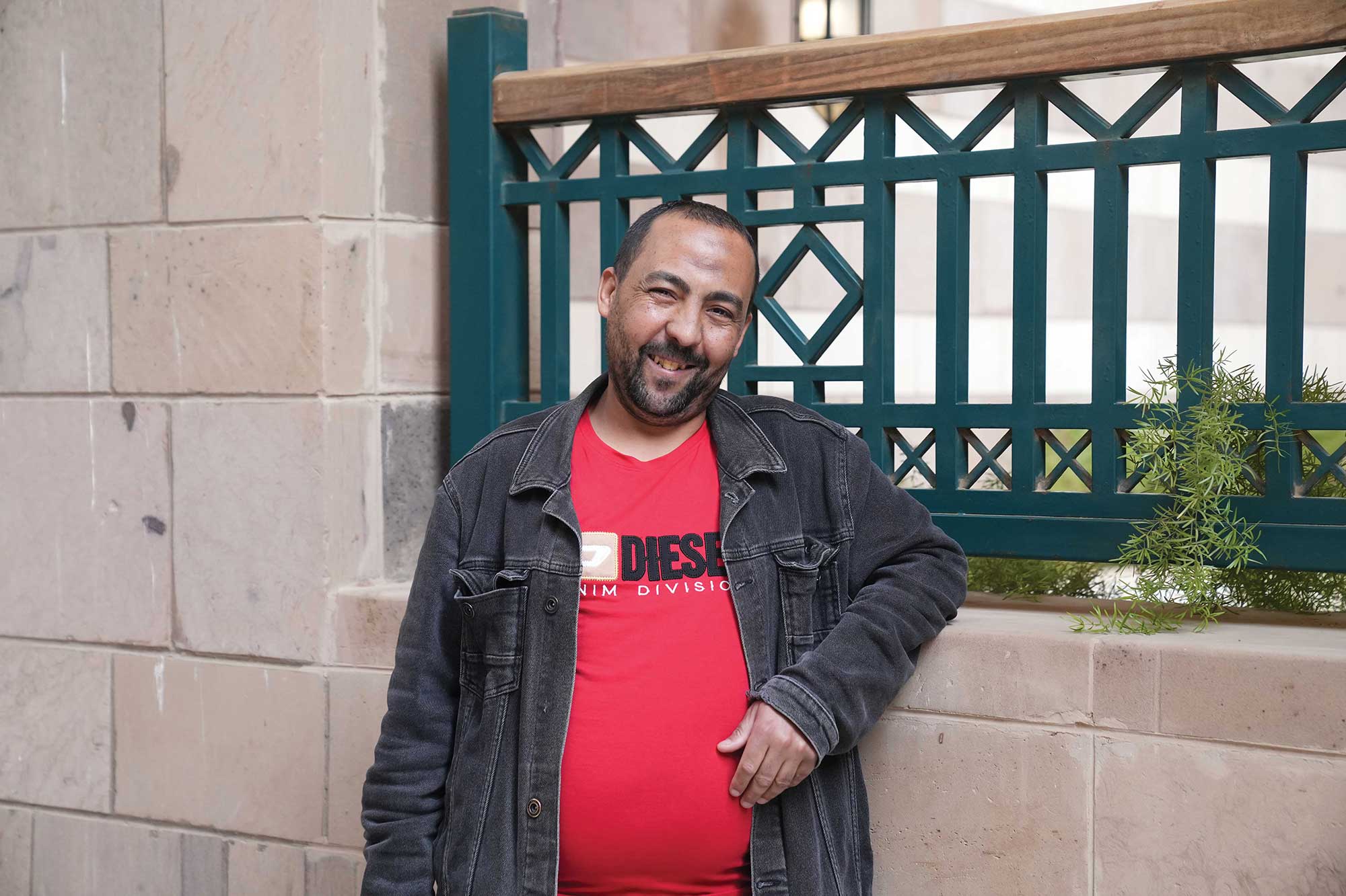 Mohamed Salah, known as Tata, stands smiling outdoors on AUC’s campus, leaning against a low wall, wearing a red T-shirt and dark jacket.
