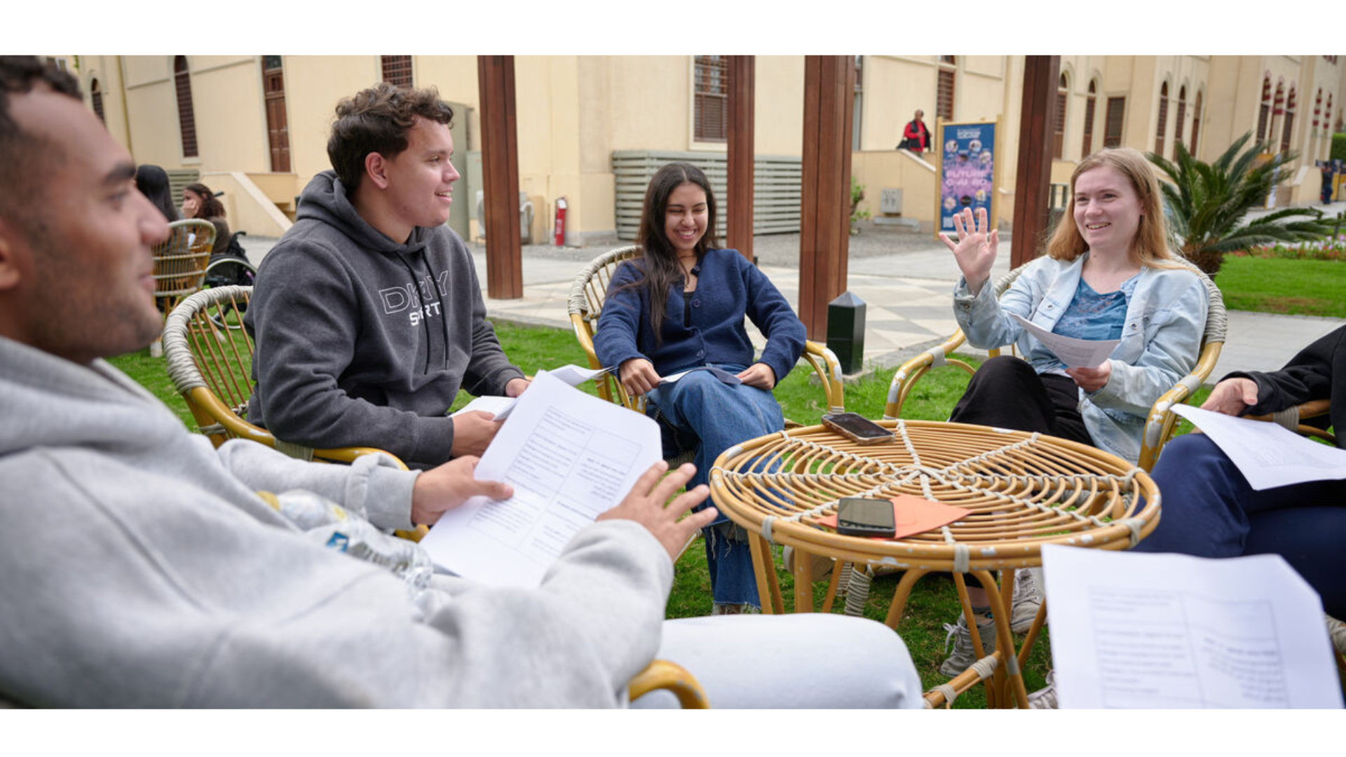 Students hold paper pamphlets while seated in chairs outside and talk with each other
