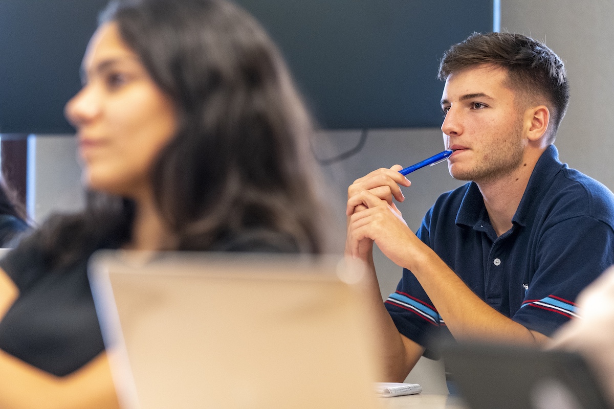 A student listens thoughtfully during a class discussion while holding a pen.