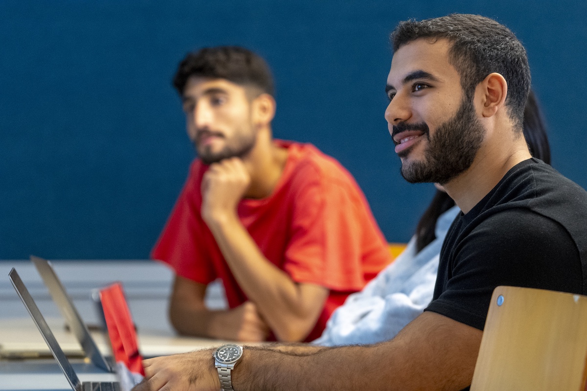 Two students sit at a table with laptops, listening attentively during a classroom discussion while one smiles and looks toward the speaker.