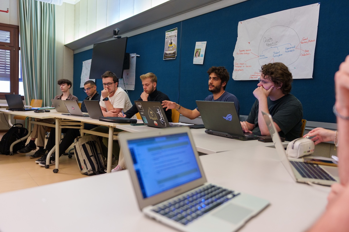 Students sit around tables with laptops during a collaborative classroom discussion.