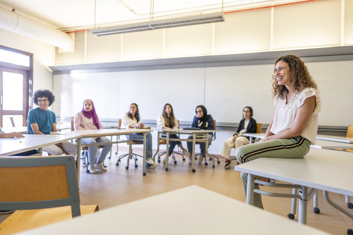 An instructor sits at the front of a classroom speaking with students seated in a circle during a discussion.