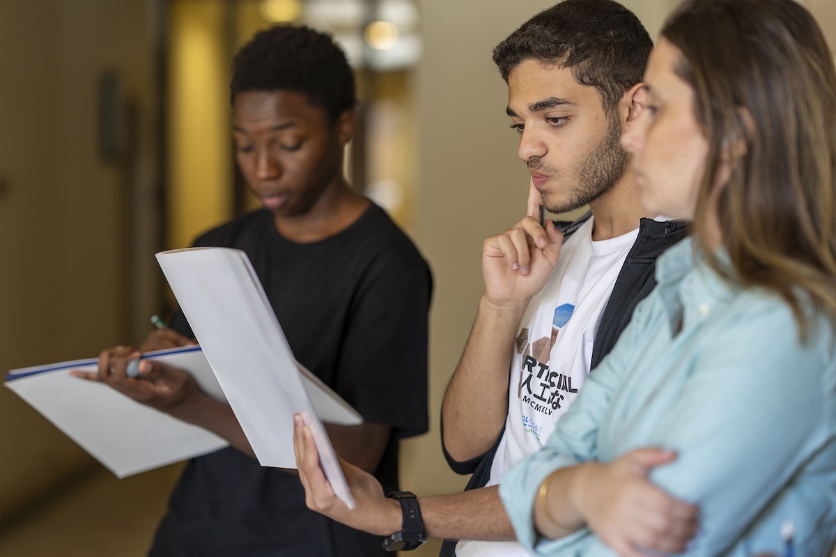 Students review printed documents together while discussing ideas in a hallway.